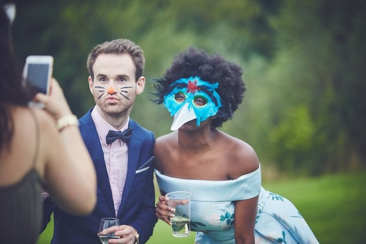 Woman taking photo of two people, a man with painted cat whiskers and a woman with a bird mask