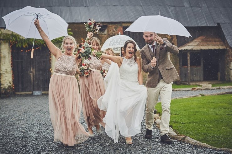 Bride and groom with bridesmaids holding umbrellas celebrating in the rain